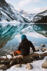Back view of anonymous tourist in sweater and hat raising arms and stretching while sitting on coast of Lake Louise against snowy mountain ridge on winter day in Alberta, Canada — Stock Photo