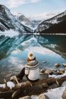 Vista trasera de la turista anónima pensativa en suéter y sombrero sentado en la costa del lago Louise contra la cresta de montaña nevada en el día de invierno en Alberta, Canadá - foto de stock