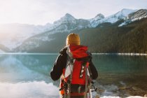 Back view of anonymous hiker with backpack walking against calm Lake Louise and snowy mountains in sunny winter morning in Banff National Park — Stock Photo