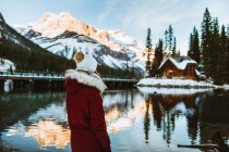Visão traseira de turista feminina irreconhecível em outerwear admirando barraca de madeira e cume de montanha enquanto estava na costa nevada do Lago Esmeralda no dia de inverno na Colúmbia Britânica, Canadá — Fotografia de Stock