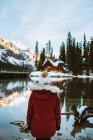 Vue arrière d'une touriste féminine méconnaissable en vêtements de dessus admirant une cabane en bois et une crête de montagne alors qu'elle se tenait debout sur la côte enneigée du lac Emerald lors d'une journée d'hiver en Colombie-Britannique, Canada — Photo de stock