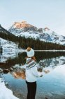 Mujer con ropa de abrigo de pie en madera a la deriva cerca de aguas tranquilas de Emerald Lake contra el paseo por la montaña nevada y el bosque de coníferas en el día de invierno en Columbia Británica, Canadá - foto de stock