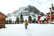 Visão traseira de uma mulher na moda irreconhecível andando em exposição perto da cabana de madeira na costa do Lago Bow no dia de inverno em Alberta, Canadá — Fotografia de Stock