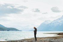 Side view of female traveler browsing cellphone while standing on shore of Abraham Lake on cloudy day in Alberta, Canada — Stock Photo