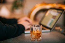 Crop anonimo persona digitando su netbook seduto a tavola con bicchiere di whisky servito con fetta di arancia — Foto stock