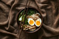 Top view of ceramic bowl with delicious ramen and chopsticks placed on table covered with brown stablecloth — стоковое фото