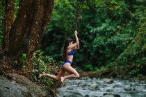 Seitenansicht einer jungen Abenteurerin in Badebekleidung, die während ihres Sommerurlaubs in der Provinz Alajuela in Costa Rica über einen Bach springt, der durch üppigen Regenwald fließt — Stockfoto