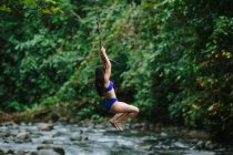 Seitenansicht einer jungen Abenteurerin in Badebekleidung, die während ihres Sommerurlaubs in der Provinz Alajuela in Costa Rica über einen Bach springt, der durch üppigen Regenwald fließt — Stockfoto