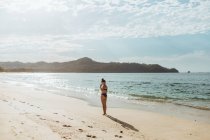 Ganzkörper junge Frau im Bikini steht allein am leeren Sandstrand in der Nähe des winkenden Ozeans und genießt den sonnigen Tag im Urlaub in Tamarindo Costa Rica — Stockfoto
