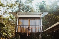 Снизу полное тело молодой длинноволосой женщины в стильных купальниках, сидящей на балконе пляжного дома, расположенного рядом с зелеными деревьями в летний день в городе Увита в Коста-Рике — стоковое фото