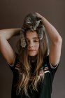 Мечтательная девочка-подросток с пушистым милым котом на голове на коричневом фоне в студии — стоковое фото