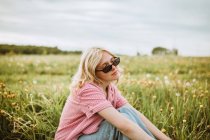 Side view of young serene female in trendy outfit sitting in blossoming meadow in summer and looking away — Stock Photo