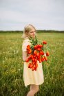 Vue latérale du contenu femelle en robe debout avec un bouquet de fleurs de tulipes rouges dans la prairie en été et regardant loin — Photo de stock