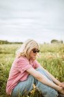 Vista laterale della giovane donna serena in abito alla moda seduta nel prato fiorito in estate e guardando altrove — Foto stock
