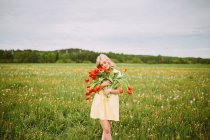 Contenido femenino en vestido de pie con ramo de flores de tulipán rojo en el prado en verano con los ojos cerrados - foto de stock