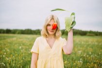 Delicate female covering eye with red tulip flower while standing in field and looking at camera — Stock Photo