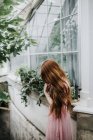 Dreamy unrecognizable redhead female in dress standing near glass windows of glasshouse with lush plants — Stock Photo