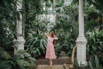 Full body of redhead female standing on tiptoes with hands on head among tropical palm and plants in glasshouse — Stock Photo
