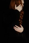 Back view of unrecognizable crop tender boyfriend tenderly embracing redhead girlfriend while standing in dark studio on black background — Stock Photo