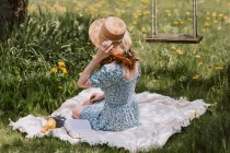 Side view of unrecognizable female in dress and straw hat sitting on picnic blanket on green meadow near swings in summer countryside — Stock Photo