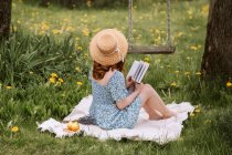 Вид збоку на невпізнавану жінку в сукні і солом'яному капелюсі читає роман, сидячи на пікніку на зеленому лузі біля гойдалок в літній сільській місцевості — стокове фото