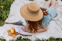 Alto ângulo de fêmea irreconhecível em vestido com chapéu de palha no rosto deitado com livro aberto em manta de piquenique enquanto relaxa sozinho e desfruta de fim de semana de verão no campo — Fotografia de Stock