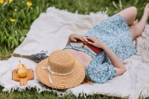 Высокий угол неузнаваемой женщины в платье с соломенной шляпой на лице, лежащей с открытой книгой на одеяле для пикника в одиночестве и наслаждающейся летними выходными в сельской местности — стоковое фото