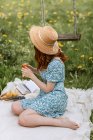 Боковой вид на неузнаваемую молодую босиком женщину в сарафане и соломенной шляпе, расслабляющую стакан напитка на одеяле для пикника с открытой книгой, наслаждаясь летним днем в одиночестве в сельской местности — стоковое фото