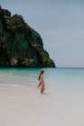 Retrovisore corpo completo di donna senza volto in abito in piedi sulla spiaggia di sabbia vicino all'acqua azzurra contro scogliere rocciose in Thailandia — Foto stock