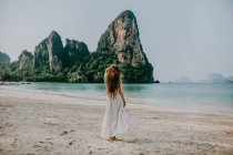 Вид сзади полное тело безликой женщины в белом платье, стоящей на песчаном пляже возле лазурной воды против скалистых скал в Таиланде — стоковое фото