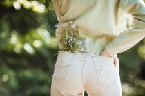Вид ззаду обрізаної дівчини-підлітка з польовими квітами в джинсовій кишені, що стоїть в літньому парку — стокове фото
