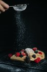 Crop anonymous chef sprinkling sugar powder on sweet waffles served with fresh berries on black background — Stock Photo