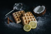 Alto ângulo de waffles vegan saborosos servidos na mesa preta com fatias de coco e limão — Fotografia de Stock