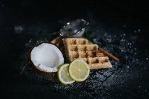 Высокий угол вкусных веганских вафель подается на черном столе с кокосовыми и лимонными ломтиками — стоковое фото