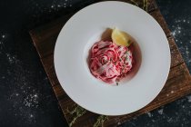 Vista superior de la apetitosa pasta de remolacha rosa vegana con rodaja de limón y ramita de tomillo servida sobre tabla de madera sobre mesa negra - foto de stock