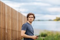 Вид збоку чоловічий спортсмен слухає музику в навушниках, стоячи біля озера під час тренувань — стокове фото