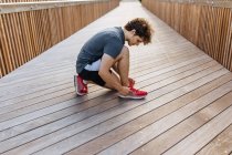 Вид сбоку на здорового бегуна, завязывающего шнурки на кроссовках на деревянной набережной — стоковое фото
