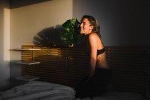 Sesión de fotos de una sensual mujer bonita sonriendo entre la luz y las sombras en lencería en la cama mirando hacia otro lado - foto de stock