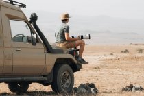 Side view of male traveling photographer sitting on offroader and taking picture on camera with telephoto lens during safari in savannah in summer — Stock Photo