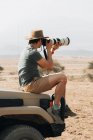 Vista lateral del fotógrafo viajero masculino sentado en offroader y tomando fotos en cámara con teleobjetivo durante el safari en Savannah en verano - foto de stock