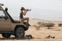 Вид сбоку на путешествующего фотографа-мужчину, сидящего на внедорожнике и фотографирующего на камеру с объективом телефона во время сафари в саванне летом — стоковое фото