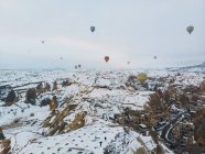 Vue imprenable sur drone de montgolfières colorées volant au-dessus de vieux villages ouchisars et de terrains rocheux enneigés avec des spectateurs lors d'une froide journée d'hiver en Cappadoce, Turquie — Photo de stock