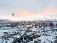 Increíble vista de drones de coloridos globos de aire caliente volando sobre el antiguo asentamiento de Uchisar y el terreno rocoso nevado con los espectadores en el frío día de invierno en Capadocia, Turquía - foto de stock