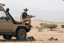 Side view of male traveling photographer sitting on offroader and taking picture on camera with telephoto lens during safari in savannah in summer — Stock Photo