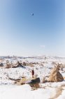 Corps entier méconnaissable touriste femelle debout sur la pierre et admirant les montgolfières volant dans un ciel bleu sans nuages sur un terrain montagneux enneigé par une journée ensoleillée d'hiver en Cappadoce, Turquie — Photo de stock