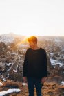 Young male in casual outfit looking away while standing against old Uchisar settlement covered with snow and sunset sky in winter evening in Cappadocia, Turkey — Stock Photo