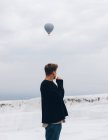 Путешествуя неузнаваемым человеком в повседневной одежде, смотрящим в сторону, стоя на белом холме минерального образования против сельской местности на горизонте и воздушном шаре, летящем в сером небе в Турции — стоковое фото