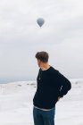 Подорожуючий незнайомий чоловік у звичайному одязі, що дивиться через плече, стоячи на білому пагорбі з мінеральними формами проти сільської місцевості за обрієм і повітряною кулею, що літає у сірому небі в Туреччині. — стокове фото