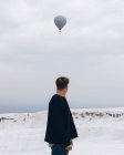Подорожуючий незнайомий чоловік у повсякденному одязі, який відводить погляд убік, стоячи на білому пагорбі мінеральних споруд проти сільської місцевості за обрієм і повітряною кулею, що літає на сірому небі в Туреччині. — стокове фото