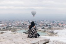 Back view of unrecognizable melancholic female in warm clothes sitting on stone slabs and embracing self while watching for big gray air balloon racing in cloudy sky over foggy city in overcast cool weather in Turkey — Stock Photo